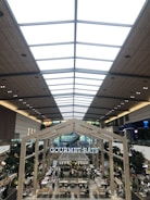 A spacious and modern indoor food court with high ceilings featuring a skylight and wooden paneling. The area is bustling with people dining at tables. Large trees are placed strategically around the area, adding greenery. A prominent sign reads 'GOURMET EATS' above the seating area.