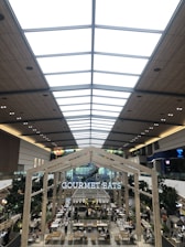 A spacious and modern indoor food court with high ceilings featuring a skylight and wooden paneling. The area is bustling with people dining at tables. Large trees are placed strategically around the area, adding greenery. A prominent sign reads 'GOURMET EATS' above the seating area.