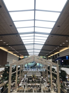 A spacious and modern indoor food court with high ceilings featuring a skylight and wooden paneling. The area is bustling with people dining at tables. Large trees are placed strategically around the area, adding greenery. A prominent sign reads 'GOURMET EATS' above the seating area.