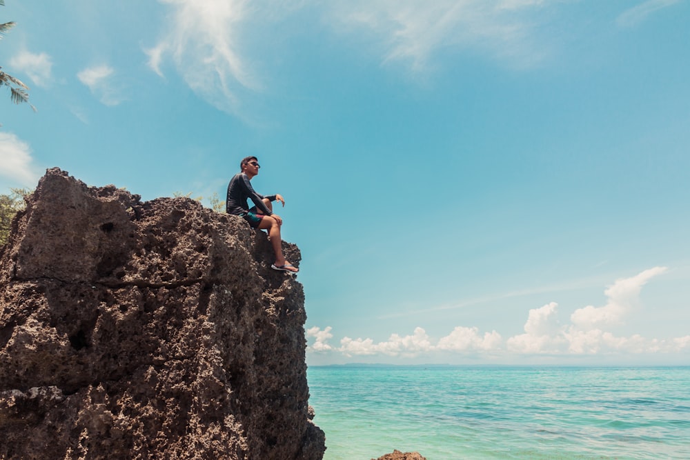man in blue t-shirt sitting on rock formation near sea during daytime
