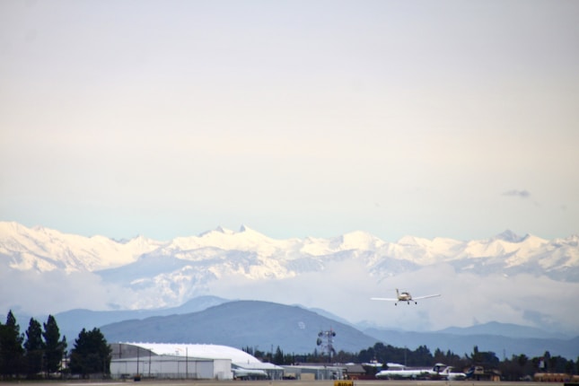 A small airplane is flying over an airstrip with snowy mountains in the background. The scene includes hangars and other airport infrastructure at the base. The landscape features trees along with sprawling mountains covered in snow.
