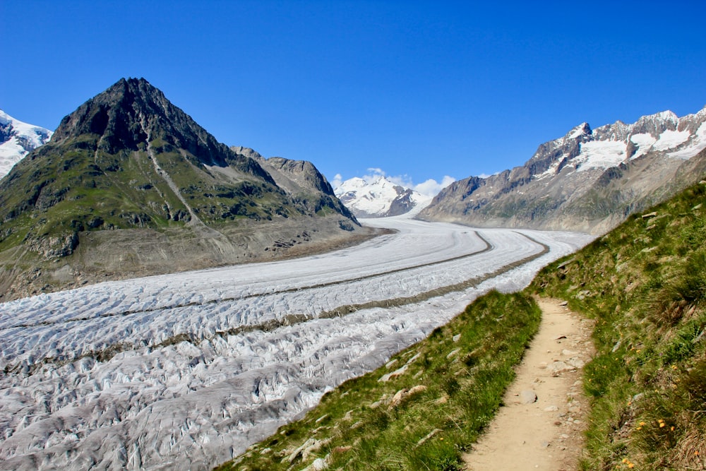 gray road between green grass field near mountain under blue sky during daytime