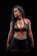 A woman with curly black hair is wearing a dark athletic outfit, including a sports bra and shorts, and holding a kettlebell. The background is completely black, emphasizing her muscular physique.