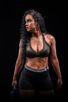 A woman with curly black hair is wearing a dark athletic outfit, including a sports bra and shorts, and holding a kettlebell. The background is completely black, emphasizing her muscular physique.