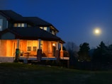 A two-story house with illuminated windows and porch lights against a dark blue night sky. The moon is visible in the right side of the image, casting a serene glow. Surrounding the house are trees and a grassy lawn.
