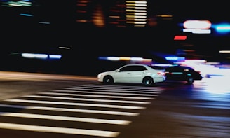 Two cars are speeding through a city intersection at night, with blurred motion indicating fast movement. Bright lights from street lamps and blurred trails from the car lights create a dynamic and energetic scene. The crosswalk is illuminated by the streetlights, contrasting sharply with the dark surroundings on the sides.