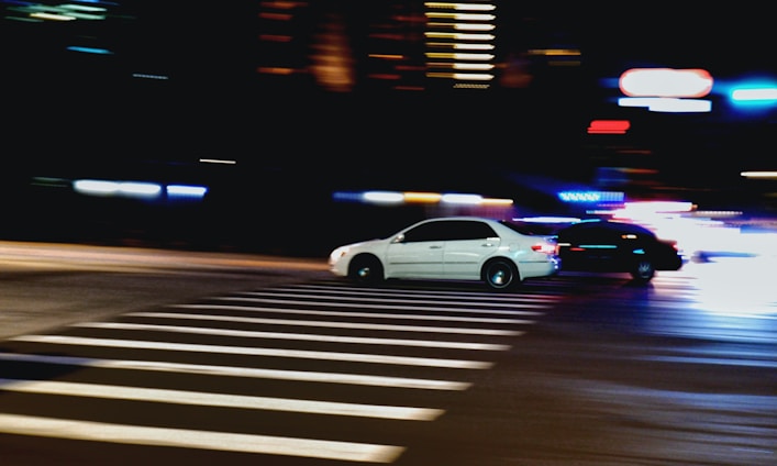 Two cars are speeding through a city intersection at night, with blurred motion indicating fast movement. Bright lights from street lamps and blurred trails from the car lights create a dynamic and energetic scene. The crosswalk is illuminated by the streetlights, contrasting sharply with the dark surroundings on the sides.