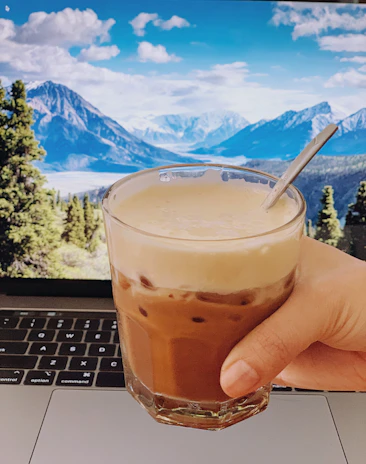 A close-up of a person’s hand holding a steaming cup of coffee next to a laptop showing weather maps.