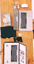Two open laptops positioned on a wooden table alongside a mouse, some notebooks, a smartphone, and glasses of iced coffee and lemon water. The laptops display different content on their screens, surrounded by casual items, suggesting a collaborative or study setup.
