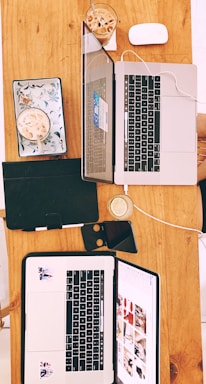 Two open laptops positioned on a wooden table alongside a mouse, some notebooks, a smartphone, and glasses of iced coffee and lemon water. The laptops display different content on their screens, surrounded by casual items, suggesting a collaborative or study setup.