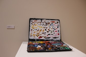 An open laptop with its screen and keyboard covered in varieties of paint and brush strokes sits on a white pedestal. The paint is applied thickly and appears to mimic the arrangement of colors on an artist's palette. Some labels are seen next to the piece, indicating it is part of an exhibit.