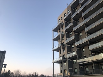 A modern, multi-story apartment building with numerous balconies is set against a clear blue sky. The structure is predominantly made of concrete with glass railings. In the background, there are bare trees and a section of another building.