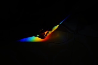 A hand is partially visible, interacting with a beam of rainbow-colored light against a dark background. The colors of the light are vivid, ranging from blue to red across the visible spectrum.