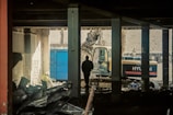 A construction site with a silhouetted person standing inside a partially demolished building. An excavator is visible in the background, and debris is scattered throughout the scene. The lighting is dim, creating a moody atmosphere.