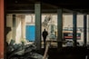 A construction site with a silhouetted person standing inside a partially demolished building. An excavator is visible in the background, and debris is scattered throughout the scene. The lighting is dim, creating a moody atmosphere.