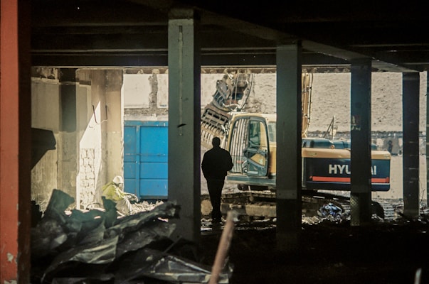 A construction site with a silhouetted person standing inside a partially demolished building. An excavator is visible in the background, and debris is scattered throughout the scene. The lighting is dim, creating a moody atmosphere.