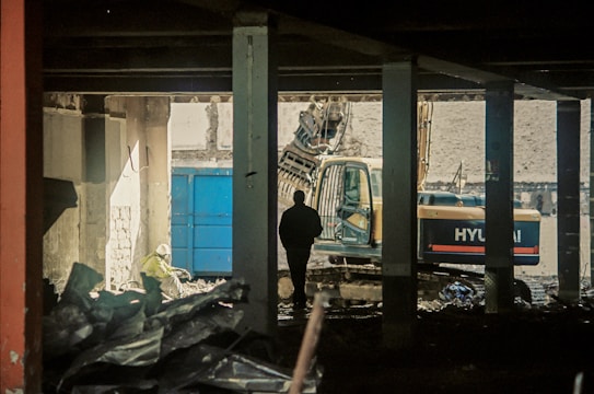 A construction site with a silhouetted person standing inside a partially demolished building. An excavator is visible in the background, and debris is scattered throughout the scene. The lighting is dim, creating a moody atmosphere.