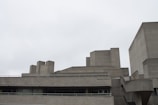 A concrete building with a brutalist architectural style. The structure features sharp angles and block-like elements, with multiple levels and flat surfaces. The sky is overcast, contributing to a muted and somber atmosphere.
