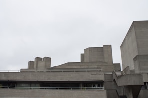 A concrete building with a brutalist architectural style. The structure features sharp angles and block-like elements, with multiple levels and flat surfaces. The sky is overcast, contributing to a muted and somber atmosphere.