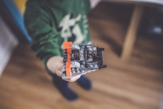 A child is holding a small LEGO creation, primarily featuring gray, orange, and black pieces. The focus is on the LEGO structure, with the child's green sweater visible but slightly out of focus in the background.