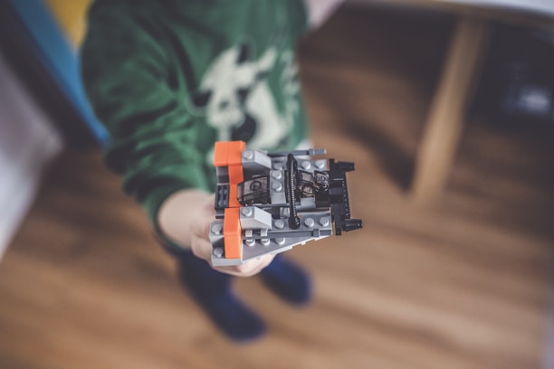 A child is holding a small LEGO creation, primarily featuring gray, orange, and black pieces. The focus is on the LEGO structure, with the child's green sweater visible but slightly out of focus in the background.