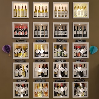 A wall display features various bottles of wine organized into individual illuminated cubbyholes. Each section has a handwritten label indicating categories such as Chardonnay, Bubbles, and Napa. Decorative elements include small paper ornaments.