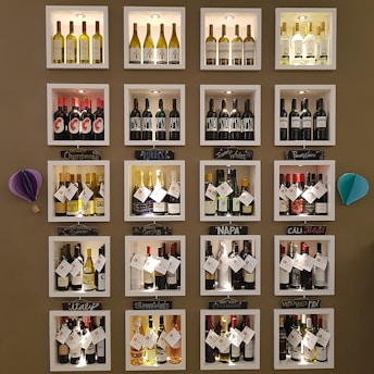 A wall display features various bottles of wine organized into individual illuminated cubbyholes. Each section has a handwritten label indicating categories such as Chardonnay, Bubbles, and Napa. Decorative elements include small paper ornaments.