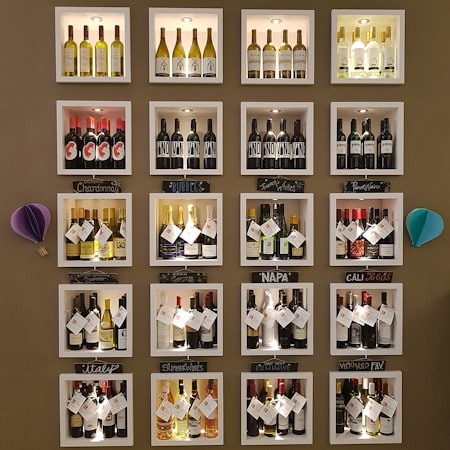 A wall display features various bottles of wine organized into individual illuminated cubbyholes. Each section has a handwritten label indicating categories such as Chardonnay, Bubbles, and Napa. Decorative elements include small paper ornaments.