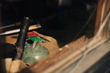 A variety of tools and objects are placed on a wooden surface, dimly lit by sunlight filtering through a window. Among the items is a green, textured ceramic piece with several holes and a handle, possibly a teapot lid or incense holder. Nearby are a wooden mallet and several small, colorful tools or utensils.