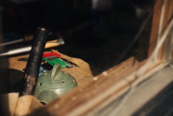 A variety of tools and objects are placed on a wooden surface, dimly lit by sunlight filtering through a window. Among the items is a green, textured ceramic piece with several holes and a handle, possibly a teapot lid or incense holder. Nearby are a wooden mallet and several small, colorful tools or utensils.