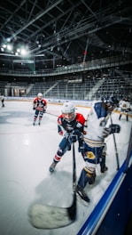 A fast-paced ice hockey game taking place in a large indoor arena. Two players are actively pursuing the puck, one wearing a red jersey with black accents and the other in a white jersey with blue stripes. The ice is smooth and shiny, reflecting the arena lights, and the stands are mostly empty, giving an impression of a private or practice session.