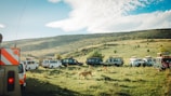 A group of safari vehicles is gathered on a grassy plain as several lions walk around them. The setting is a wide open landscape with a gentle hill in the background. The sky is partly cloudy with patches of blue.