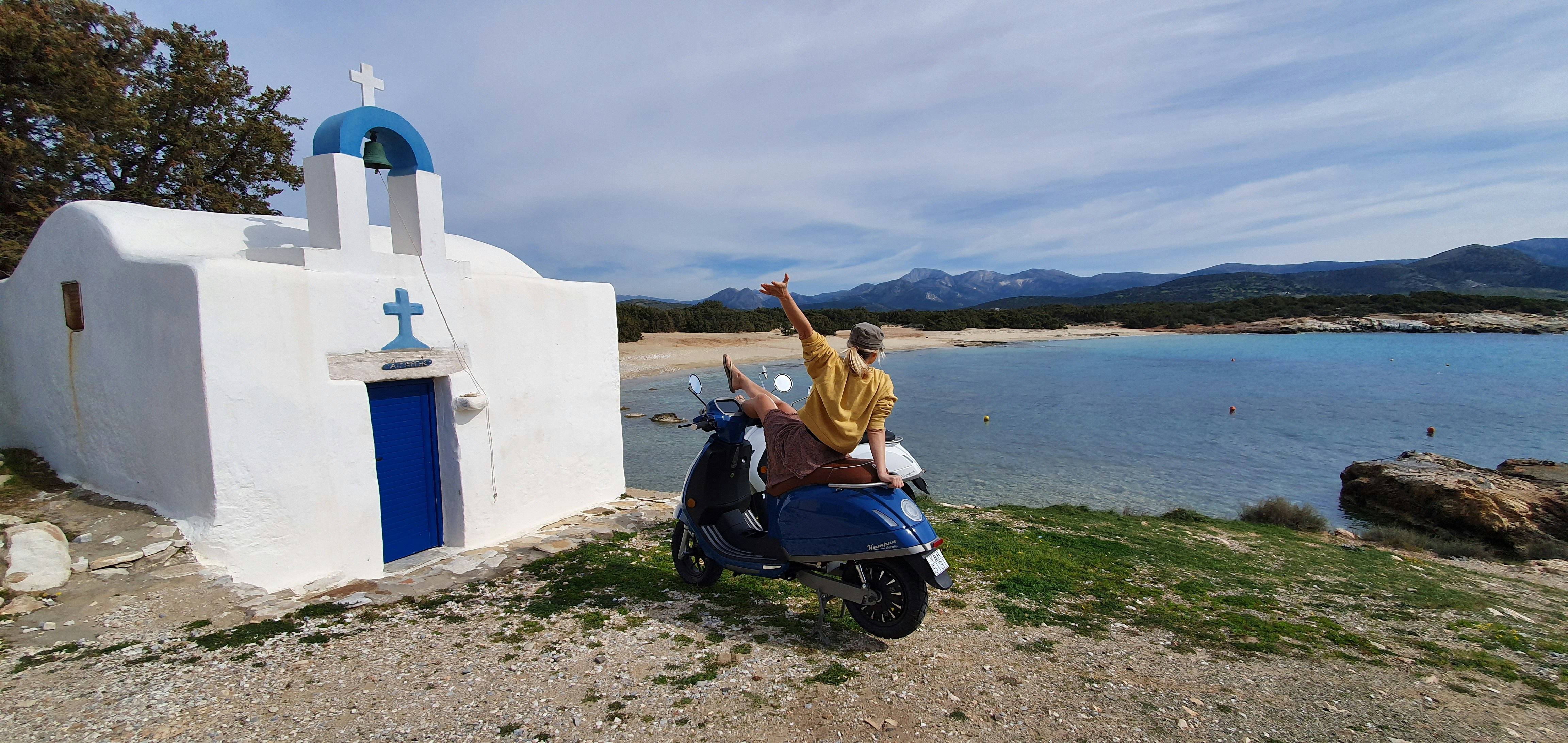 woman in brown jacket riding blue motor scooter on beach during daytime, Enjoy the view and temperatures in Greece!</p><p>A girl sitting on her electric scooter Kumpan 1954 Ri. 