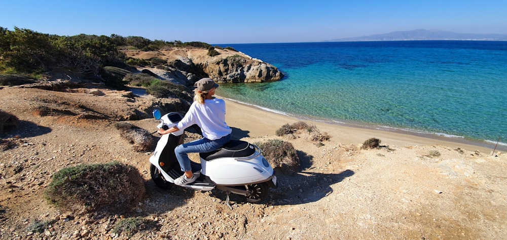 man in white shirt and blue pants sitting on white and black motor scooter on beach