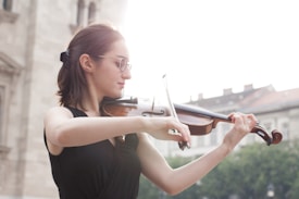 A person with glasses plays the violin outdoors with a background of blurred buildings and greenery. The person wears a black sleeveless top and appears focused on playing the instrument.