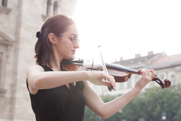 A person with glasses plays the violin outdoors with a background of blurred buildings and greenery. The person wears a black sleeveless top and appears focused on playing the instrument.