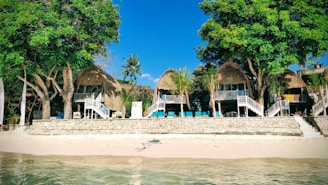 Several thatched-roof huts are situated on a sandy beach, surrounded by lush green trees. The structures are elevated with wooden staircases leading up to them. The ocean is visible in the foreground, indicating a tropical setting.