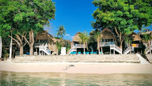 Several thatched-roof huts are situated on a sandy beach, surrounded by lush green trees. The structures are elevated with wooden staircases leading up to them. The ocean is visible in the foreground, indicating a tropical setting.