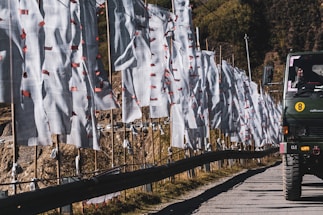 A line of white prayer flags with red tassels is displayed along a roadside, extending into the distance. A military vehicle is visible on the road, with a person inside the vehicle. The background shows rugged terrain with hills and a few scattered trees.