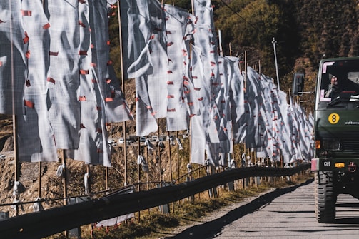 A line of white prayer flags with red tassels is displayed along a roadside, extending into the distance. A military vehicle is visible on the road, with a person inside the vehicle. The background shows rugged terrain with hills and a few scattered trees.