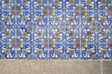 A detailed pattern of ceramic tiles featuring intricate blue and yellow floral designs against a white background. The tiles are arranged in a repetitive geometric pattern. Below the tiles, there is a stone or concrete surface.