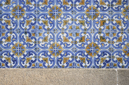 A detailed pattern of ceramic tiles featuring intricate blue and yellow floral designs against a white background. The tiles are arranged in a repetitive geometric pattern. Below the tiles, there is a stone or concrete surface.