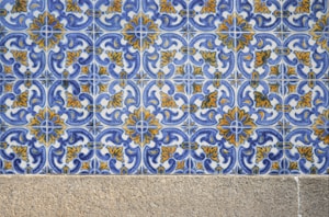 A detailed pattern of ceramic tiles featuring intricate blue and yellow floral designs against a white background. The tiles are arranged in a repetitive geometric pattern. Below the tiles, there is a stone or concrete surface.
