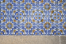 A detailed pattern of ceramic tiles featuring intricate blue and yellow floral designs against a white background. The tiles are arranged in a repetitive geometric pattern. Below the tiles, there is a stone or concrete surface.