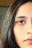 A close-up of the right side of a person's face, focusing on one eye, part of the nose, and the mouth. The person has long, straight dark hair and natural skin tone with visible textures and light reflection. The lighting is bright and even, highlighting facial features.