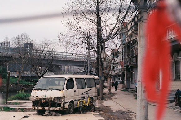 An urban street scene featuring a rundown van with a missing front bumper and an overturned bicycle lying next to it. The background includes leafless trees, residential buildings, and an elevated highway. A blurred red object is visible in the foreground, while a few pedestrians walk in the distance.