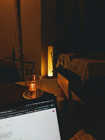 A dimly lit setting with a candle placed on a table beside a laptop showing a web page. The background includes a table with a tablecloth and a softly glowing floor lamp.