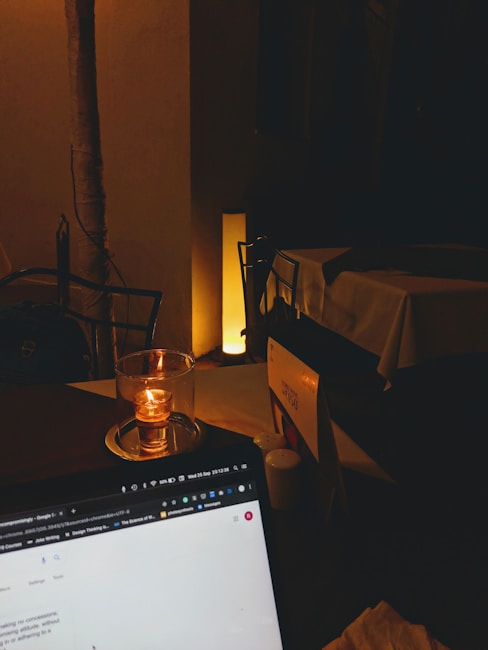 A dimly lit setting with a candle placed on a table beside a laptop showing a web page. The background includes a table with a tablecloth and a softly glowing floor lamp.
