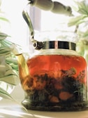 A glass teapot filled with a richly colored herbal tea is placed among green, leafy plants. The tea is transparent and has visible slices of lemons and herbal leaves floating inside. Sunlight streams through a window, casting soft, warm light and shadows around the scene, enhancing the natural and relaxing atmosphere.