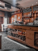 A cozy coffee shop interior featuring wooden decor, a counter with coffee brewing equipment, shelves stocked with jars and bottles, and signs displaying coffee-related messages. Ambient lighting with small string lights adds warmth to the atmosphere.